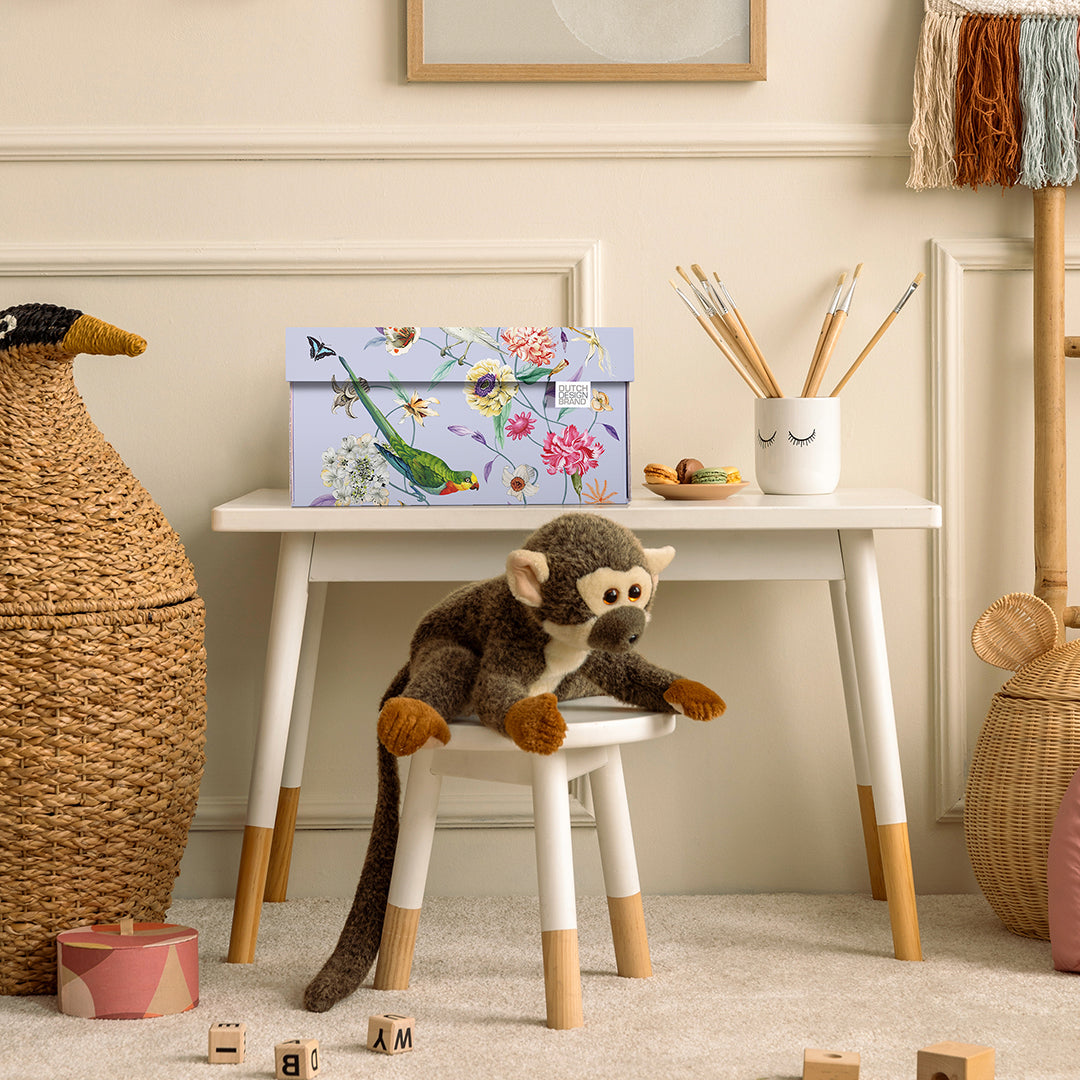 Lila opbergdoos met kleurrijk bloemen- en vogelmotief op een wit kinderbureau in een stijlvol ingerichte kinderkamer.