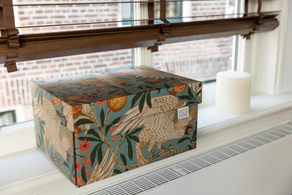 A storage box with a vibrant print of cockatoos and pomegranates. The box features detailed illustrations of white cockatoos with yellow crests among green leaves and red pomegranates on a soft blue background. The artwork has a botanical and tropical aesthetic.
