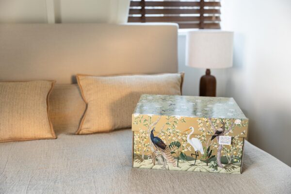 A storage box adorned with the "Golden Paradise" design, showcasing a variety of birds such as a peacock and a white heron amidst lush flora and fauna on a gold background. The box is detailed with illustrations of blooming white flowers, lemon fruits, and dragonflies.